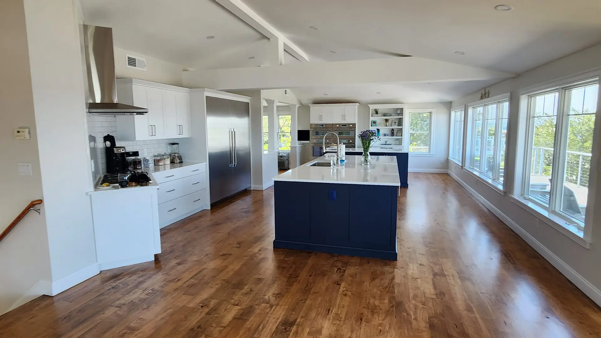 Open-concept kitchen featuring modern cabinetry, a large island with a white countertop, stainless steel appliances, and abundant natural light from large windows, highlighting a blend of contemporary design and functional living space.